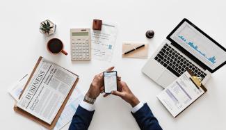 Business person with a newspaper, calculator, computer, and clipboard on desk.