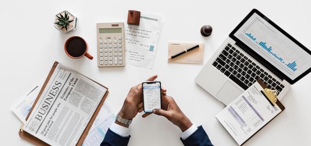 Business person with a newspaper, calculator, computer, and clipboard on desk.